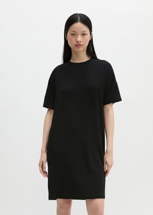 Femme aux longs cheveux noirs portant une robe noire simple, ample et à manches courtes, debout devant un fond blanc.