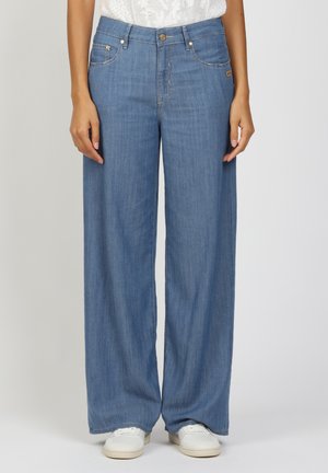 Frau trägt lockere hellblaue Jeans aus Denim und weiße Sneaker, steht vor einem einfarbigen hellen Hintergrund.