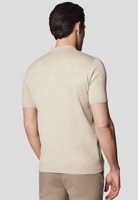 Jasno beżowa koszulka polo z krótkim rękawem o gładkiej teksturze. Posiada żebrowany kołnierzyk i dół, z dopasowanym krojem widocznym z tyłu.
