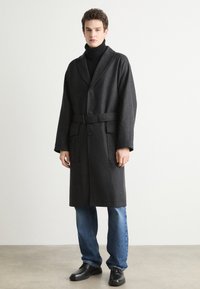 Dark gray, knee-length coat with a belt, large front pockets, and a notched collar. Worn over a black turtleneck and paired with blue jeans.