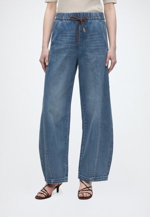Wide-leg blue denim jeans with elastic waistband and brown drawstring, worn with light top and brown strappy heeled sandals.