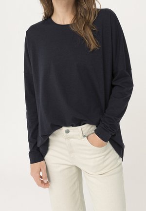 Femme portant un haut noir à manches longues ample avec un col rond et un pantalon taille haute écru, main dans la poche.