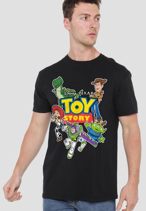 Mann trägt ein schwarzes Toy Story T-Shirt mit den Charakteren Woody, Buzz Lightyear, Jessie, Rex und Alien vor einem einfarbigen Hintergrund.