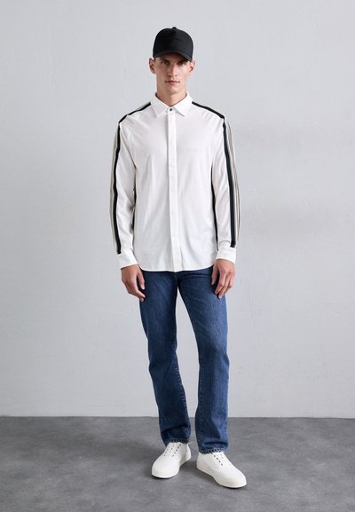 Vit långärmad skjorta med svarta sidränder, knapp-down krage, kombinerad med blå jeans och vita sneakers, bärs med en svart keps.