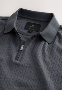 Grijze poloshirt met rits en een slanke pasvorm, met een gestructureerd visgraatpatroon. De kraag en rits hebben een bijpassende grijze tint.