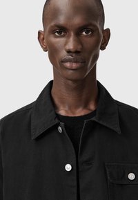 Czarna kurtka z klasycznym kołnierzem i srebrnymi napami, prezentowana na teksturowanym czarnym swetrze. Materiał jest gładki o matowym wykończeniu.