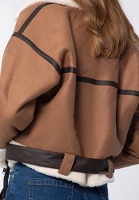 Veste en fausse suédine marron avec col en fourrure synthétique, présentant des accents en cuir noir et une taille ceinturée. Tissu texturé avec une coupe ample.