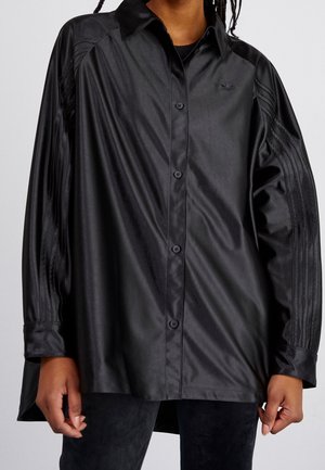 Osoba nosząca oversize'ową czarną koszulę na guziki z subtelnym logo Adidas i trzema teksturowanymi paskami na rękawach, w połączeniu z czarnymi spodniami.