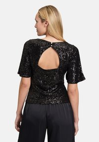 Schwarzes Paillettenoberteil mit kurzen Ärmeln, das einen runden Ausschnitt auf der Rückseite und ein Knopfdetail am Halsabschluß aufweist. Kombiniert mit fließenden schwarzen Hosen.