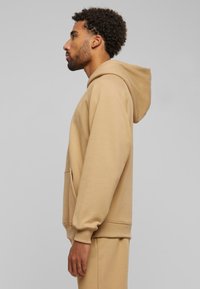 Beige hettegenser med kengurulomme, ribbede mansjetter og en myk tekstur, vist fra en sideprofil mot en ensfarget bakgrunn.