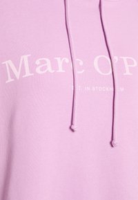 Zbliżenie na różowy materiał bluzy z kapturem z białym nadrukiem "Marc O’P" i częściowo widocznymi białymi sznurkami, które zwisają w dół.