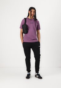 Fialové bavlnené tričko s logom, čierne jogger nohavice a čierna taška cez rameno. Čierne tenisky s bielymi pruhmi. Jednoduchý, neformálny outfit.