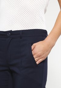 Marineblauwe broek van gladde stof, met een voorzak en een knoopsluiting. De tailleband is gestructureerd voor een getailleerde look.