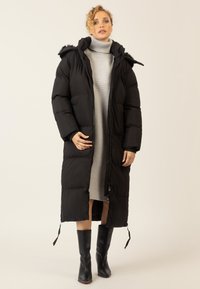 Schwarzer, gepolsterter langer Mantel mit Kapuze, strukturierten Einsätzen, Front-Reißverschluss und seitlichen Kordeln, getragen über einem grauen, gestreiften Kleid und schwarzen Stiefeln.