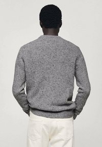 Homme de dos portant un pull en maille gris moucheté et un pantalon écru, les mains dans les poches, sur un fond uni.