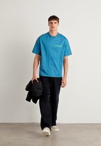 Μπλε βαμβακερό t-shirt με βραχίονες και πράσινο λογότυπο, συνδυασμένο με μαύρο παντελόνι και κρατώντας μια μαύρη ζακέτα, τοποθετημένο σε ουδέτερο φόντο.