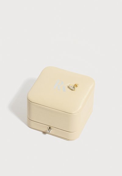 Κρεμ χρώματος κουτί κοσμημάτων με ένα μικρό χρυσό και ασημένιο σκουλαρίκι καρφί, τοποθετημένο πάνω, σε απλό λευκό φόντο.