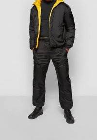 Person som bär en svart vadderad jacka med gul foder, svarta vadderade byxor, svart skjorta och svarta sneakers, stående med händerna i fickorna.