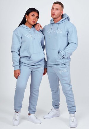 Due modelle che indossano felpe con cappuccio e pantaloni della tuta abbinati di colore azzurro chiaro, con scarpe da ginnastica bianche, posano davanti a uno sfondo semplice.