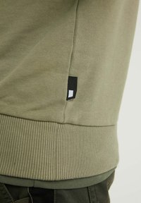 Felpa verde oliva con polsini e orlo a coste. Presenta un piccolo etichetta nera con un rettangolo bianco vicino alla cucitura laterale. Tessuto di cotone liscio.
