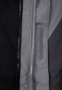 Vnitřek šedé bundy se stříbrným zipem, černou síťovinou a páskem na suchý zip. Hladká textura s různými materiály a kontrastními barvami.