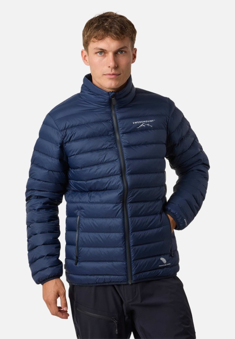 Giacca a vento blu navy con colletto alto, pannelli cuciti e due tasche laterali con zip. Presenta il logo sul lato sinistro del petto. Texture liscia.