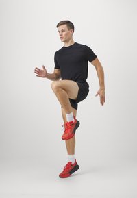 Juodas sportinis marškinėlis, juodi šortai, raudoni bėgimo bateliai su juodais padais ir raišteliais, vidurinė pozicija, balti kojinai, lygus fonas.