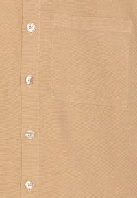 Camicia di cotone beige con una texture liscia, caratterizzata da cinque bottoni e una tasca sul petto sinistro. Design semplice con leggeri dettagli di cucitura.