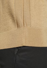 Ourlet de pull beige en tricot porté sur un pantalon en cuir noir, mettant en valeur la texture du tricot et le contraste des tissus.