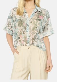 Short-sleeve, light green floral blouse with blue and pink accents, featuring a button-down front and a relaxed fit, paired with beige trousers.