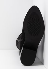 Botte noire avec semelle texturée et fermeture éclair sur le côté, positionnée debout contre un mur d'angle blanc.