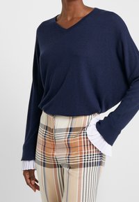 Granatowy sweter z dekoltem w serek i luźnymi rękawami. Połączony z spodniami w kratę w beżowo-czarną i pomarańczową wzory, z białymi falbaniastymi mankietami.