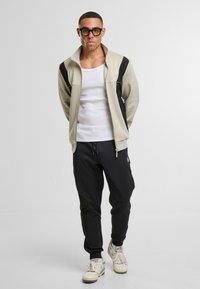 Světle béžová mikina na zip s černými akcenty, kombinovaná s černými tepláky a bílými teniskami. Model má krátké vlasy a nosí brýle.