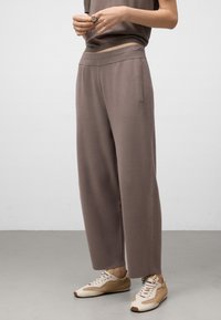 Persoon in losse taupe broek en beige sneakers, staande op een grijze vloer tegen een witte muur.