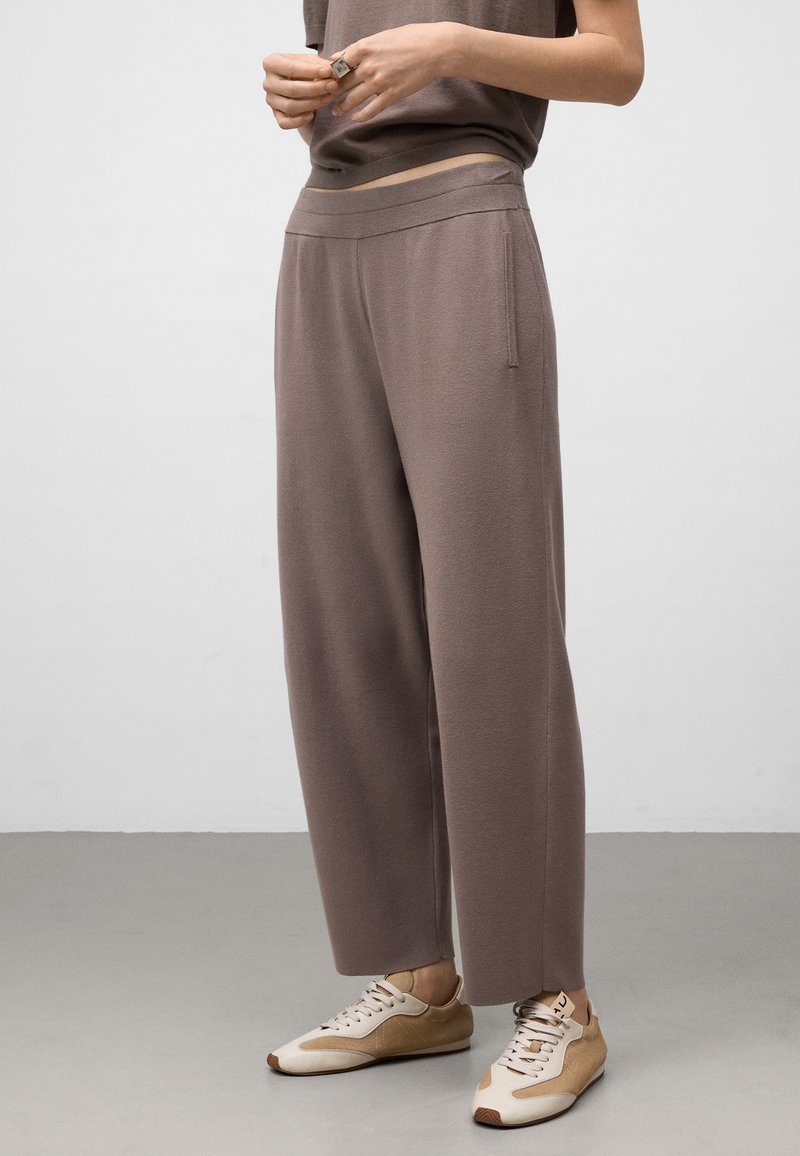 Persoon in losse taupe broek en beige sneakers, staande op een grijze vloer tegen een witte muur.