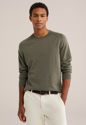Junger Mann trägt einen olivgrünen Pullover mit langen Ärmeln und weiße Hosen mit einem dunkel gewebten Gürtel, steht mit einer Hand in der Tasche.