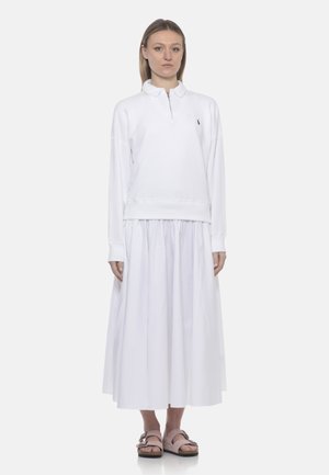 Donna in piedi che indossa una felpa bianca a maniche lunghe con mezza cerniera, una gonna midi bianca arricciata e sandali rosa con fibbia.