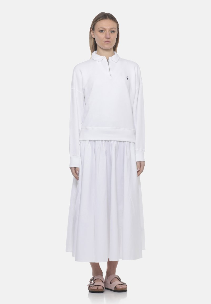 Donna in piedi che indossa una felpa bianca a maniche lunghe con mezza cerniera, una gonna midi bianca arricciata e sandali rosa con fibbia.