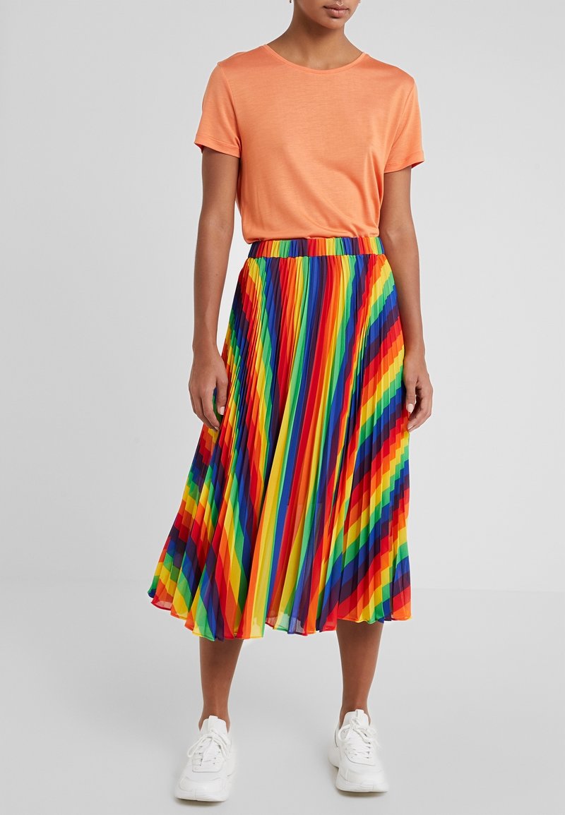 Jupe plissée arc-en-ciel avec des rayures horizontales vives, associée à un t-shirt orange à col rond. Des baskets blanches complètent la tenue.