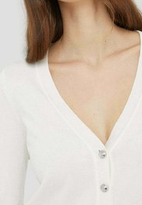 Vit kofta med djup V-ringning, tre knappar framtill och mjukt, texturerat stickat material.