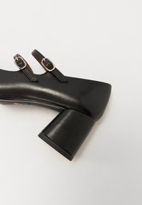 Scarpa nera con tacco alto in materiale similpelle, punta tonda, tacco spesso a blocco e due cinturini stretti con fibbie argentate.