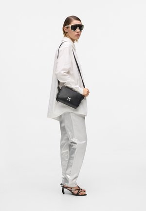 Čierna kožená kabelka cez rameno so strieborným logom, oversize biela košeľa, strieborné metalické nohavice a veľké hranaté slnečné okuliare. Sandále na podpätku.