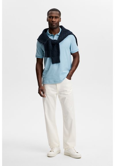 Uomo in piedi che indossa una polo azzurro chiaro, pantaloni bianchi, scarpe da ginnastica bianche, con un maglione scuro appoggiato sulle spalle e annodato davanti.