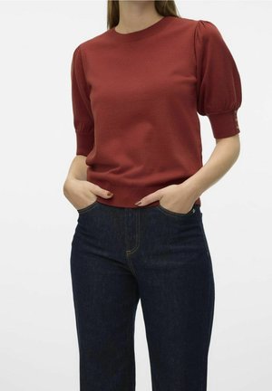 Frau trägt rostfarbenes Oberteil mit Puffärmeln und Knopfdetails und dunkelblaue Jeans, Hände in den Taschen, steht vor schlichtem Hintergrund.