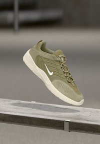 Ελιάς πράσινο αθλητικό παπούτσι Nike με λευκή σόλα και λογότυπο swoosh, που αιωρείται πάνω από μία бетонική άκρη εσωτερικού χώρου.