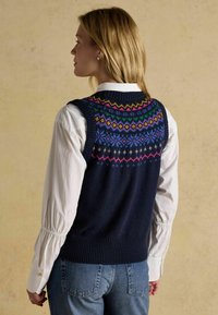 Jūras spēku adīts džemperis ar krāsainu ģeometrisku rakstu plecu daļā, uzvilkts pāri baltai krekla, ar ribotu apakšmalu un roku atverēm.