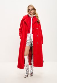 Abrigo largo de pelo sintético rojo, botas metálicas plateadas hasta la rodilla, combinado con una blusa roja, camisa blanca y pantalones cortos plateados. Textura suave, fit holgado.