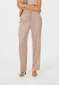 Beige satin elastic waistband trousers with a relaxed fit, featuring side pockets and a smooth, glossy texture; paired with gold-heeled sandals.