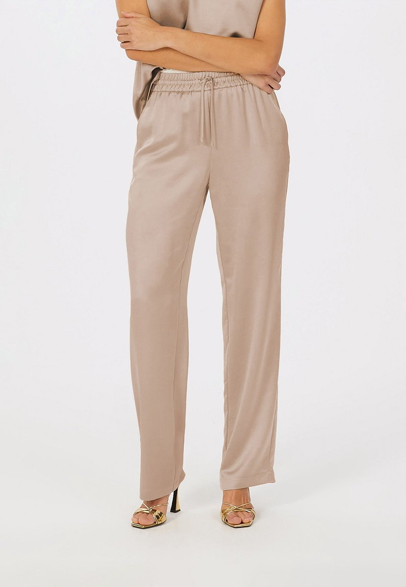 Beige satin elastic waistband trousers with a relaxed fit, featuring side pockets and a smooth, glossy texture; paired with gold-heeled sandals.