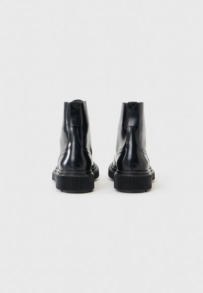Ζεύγος μαύρων γυαλιστερών δερμάτινων μποτών αστραγάλου με παχιά αυλακωτά πέλματα, από πίσω, σε απλό ανοιχτό φόντο.